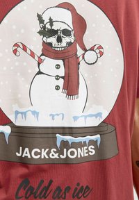 Rode shirt met een afbeelding van een sneeuwman met een schedel als hoofd, gekleed in een kerstmanmuts en sjaal, die een candy cane vasthoudt, met ijseffecten eronder.
