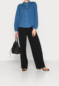 Camicia in denim con maniche a sbuffo lunghe, pantaloni neri a gamba larga e una borsa nera dalla texture particolare. Le scarpe in pelle marrone completano l'outfit.