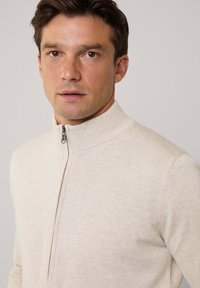 Suéter con cremallera color beige claro, con un alto cuello acanalado. Textura suave y diseño ajustado con un sutil patrón jaspeado.
