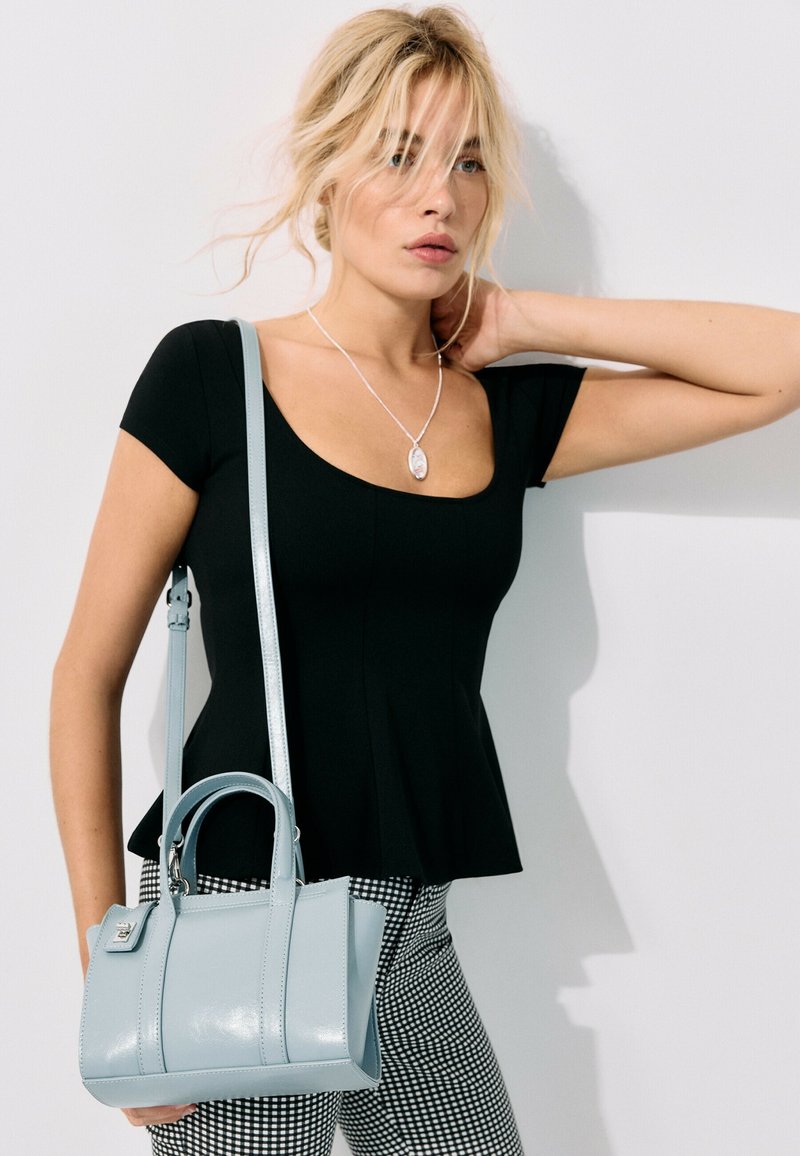 Femme blonde aux cheveux lâchés porte un haut noir, un pantalon à carreaux, un collier pendentif en argent et porte un sac à main bleu clair à l'épaule.