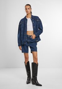 Donna con indosso una giacca di jeans scura oversize e pantaloni shorts abbinati, top corto bianco e stivali neri fino al ginocchio, in piedi con una mano in tasca.