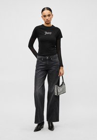 Mladá žena v čiernom viacvrstvovom tričku s dlhým rukávom a potlačou "Juicy", vyblednuté čierne široké džínsy, čierne čižmy, drží malú striebornú kabelku.