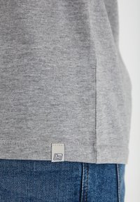 Camiseta de algodón gris con una textura sutil, abertura lateral y una etiqueta de logo en el dobladillo, combinada con jeans de mezclilla azul.