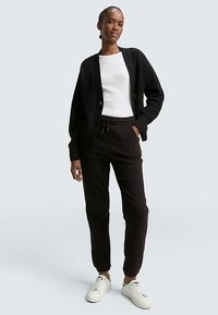 Schwarze, gerippte Strickjacke über weißem T-Shirt, kombiniert mit dunklen Jogginghosen und weißen Turnschuhen. Die Strickjacke hat Knöpfe und eine lockere Passform.