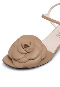 Sandalo in pelle color caramello con una parte anteriore imbottita a forma di fiore, punta aperta e sottile cinturino alla caviglia. Suola piatta con un design elegante e una superficie liscia.