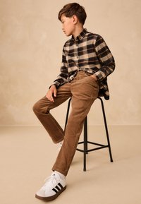 Chemise à carreaux beige et noire boutonnée, pantalon en velours côtelé marron, et baskets blanches avec des accents noirs ; assis sur un tabouret en métal noir.