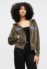 Jovem mulher com cabelo encaracolado, vestindo um casaco com capuz verde oliva com fecho e calças pretas, em pé com uma mão no bolso contra um fundo simples.