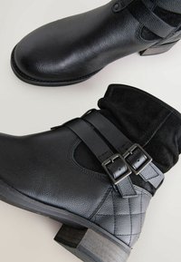 Botas negras hasta el tobillo, hechas de cuero y ante. Sus características incluyen doble hebilla, patrón acolchado y un tacón bajo apilado.