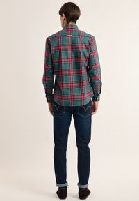 Camisa de cuadros verde y rojo con mangas largas, puños abotonados y dobladillo curvado. Combinada con jeans de mezclilla oscura y zapatos marrones.