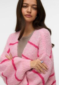 Ροζ πλεκτή ζακέτα με αντίθετες ραφές σε ματζέντα, απαλή υφή, ανοιχτό μπροστινό μέρος και χαμηλωμένους ώμους, φορεμένη πάνω από ένα μπεζ τοπ.