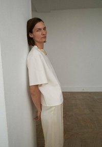 Cămașă crem cu mânecă scurtă, cu un fit lejer, asociată cu pantaloni bej deschis, fluidi. Un colier masiv din perle accentuează ținuta.