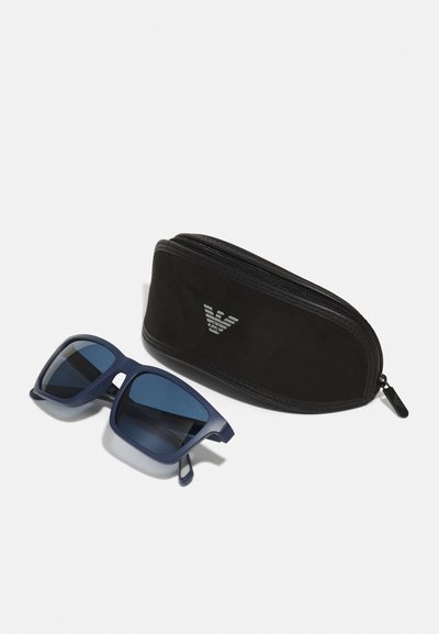 Ochelari de soare navy cu rame pătrate și lentile închise, lângă un etui negru cu fermoar ce prezintă un logo argintie. Textură mată pe rame.