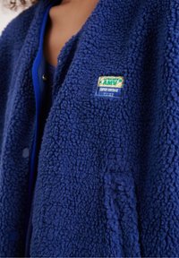Mėlynas flisinis viršutinis drabužis su faktūruota paviršiaus struktūra, apvaliu apykakliu, spaustukais ir etikete, kurioje užrašyta „LA MAISON AMY SUPER VINTAGE“.