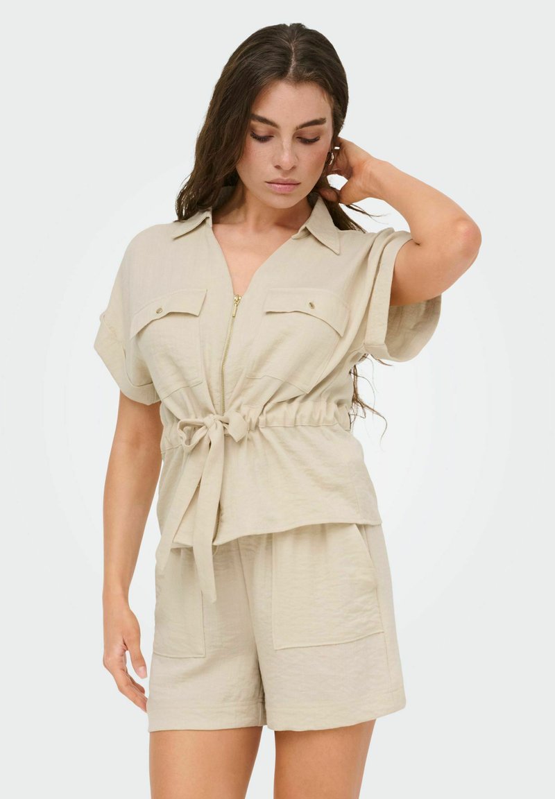 Femme en chemise beige à manches courtes zippée avec poches devant et taille nouée, short assorti avec grandes poches avant, touchant ses cheveux.