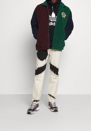 Gestructureerde fleece jas in bordeaux, navy en groen; gelaagd over een zwarte hoodie. Beige spijkerbroek met zwarte tie-dye patronen; sportieve sneakers.