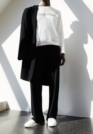 Personne portant un sweat-shirt blanc Calvin Klein, pantalon noir à jambes larges, baskets blanches et manteau noir drapé sur une épaule, debout à l'intérieur.