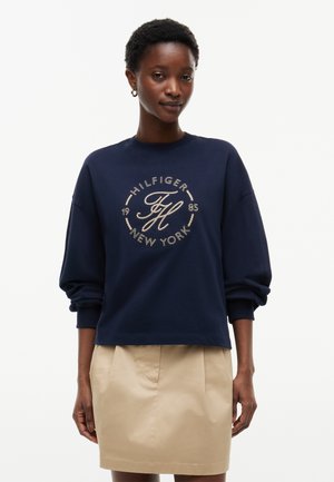 Kvinde iført en marineblå sweatshirt med teksten "Hilfiger New York" og beige nederdel, stående mod en ensfarvet hvid baggrund.