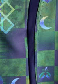 Bunt gemusterter Stoff mit grünen und blauen Motiven, darunter Rauten und Halbmondformen, kombiniert mit einem kontrastierenden dunkeln Marine-Seitenpaneel.