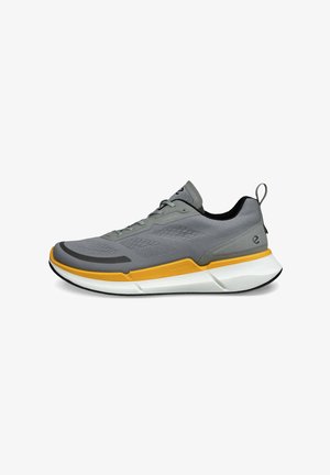 Grauer Sportschuh mit einem strukturierten Obermaterial, gelbem Akzent in der Zwischensohle, abgerundetem Zeh, schwarzem Innenraum und einer Schlaufe an der Ferse.