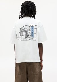 Witte oversized t-shirt met een zwarte lijntekening van een café met "Thee & Koffie" signage op de achterkant. Korte mouwen, ontspannen pasvorm.