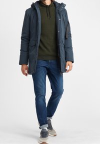 Homme portant une veste d'hiver marine sur un hoodie vert, un jean bleu à revers et des sneakers gris, marchant devant un fond blanc uni.
