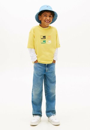 Smilende dreng iført blå spandhat, gul t-shirt med grafik over hvid langærmet trøje, blå jeans og hvide sneakers, hænderne i lommerne.