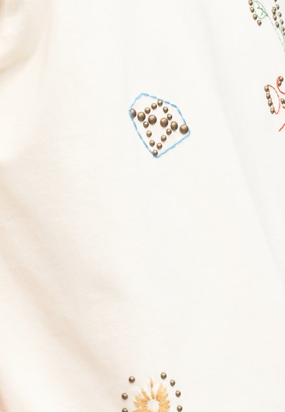 Λευκό ύφασμα με διάφορα μικρά ορειχάλκινα καρφιά διατεταγμένα σε μοτίβα, με περίγραμμα από μπλε και πορτοκαλί κεντητά ραφές.