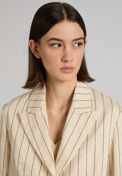 Junge Frau mit schulterlangem dunklem Haar, die einen beige gestreiften Blazer trägt und leicht nach rechts blickt vor einem schlichten Hintergrund.