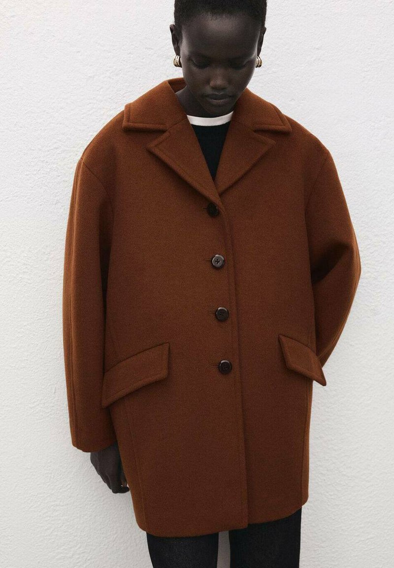 Bruine wollen jas met een vierkante gesneden vorm, reverskraag, vier knopen en klepzakken. De textuur is glad en dik, wat warmte biedt.