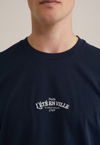 Marineblaues Baumwoll-T-Shirt mit weißem gesticktem Text, der auf der Brust "Paris l'été en ville le soleil français 1987" liest.