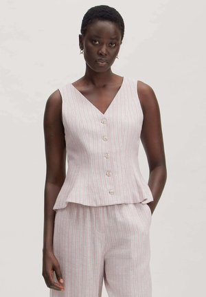 Woman with short hair wears sleeveless, buttoned, V-neck striped top and matching striped pants with hands in pockets against plain background.