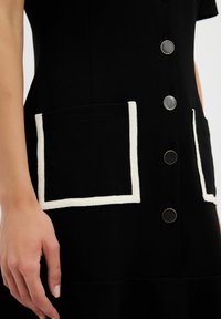 Vestido negro con dos bolsillos bordeados en blanco, botones plateados y una textura suave. Enfoque en un diseño funcional con acentos en contraste.