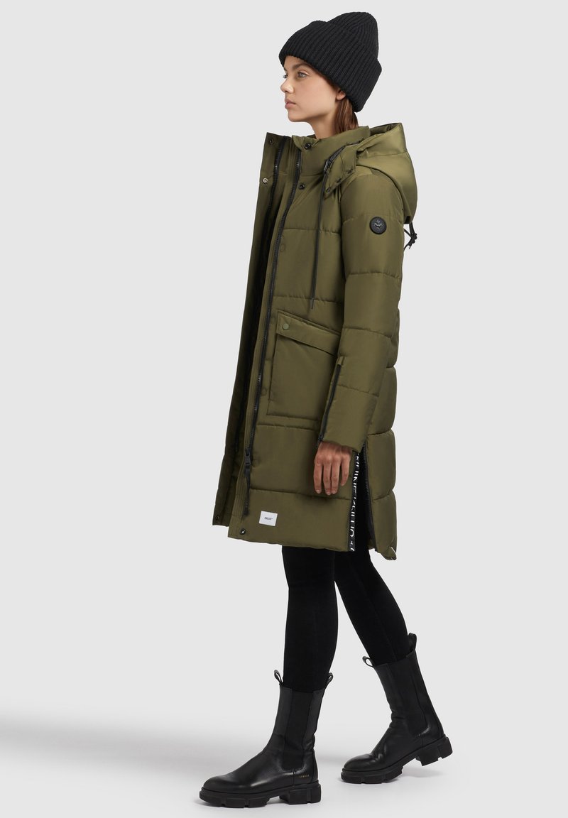 Olivengrüner langer Puffer-Mantel mit Kapuze, Reißverschluss vorne, Druckknöpfen, zwei Seitentaschen und einem schwarzen Logo-Patch am Arm. Kombiniert mit schwarzen Leggings und Stiefeln.