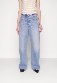 Jasnoniebieskie dżinsy o szerokim kroju z wyblakłym wykończeniem, pięcioma kieszeniami i surowym dołem. Połączone z beżowym topem w paseczki i czarnymi butami.