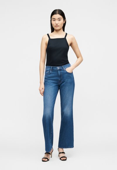 Γυναίκα στέκεται προς τα εμπρός φορώντας μαύρο τοπ με τετράγωνο ντεκολτέ, μπλε τζιν παντελόνι με ψηλή μέση και φαρδύ πόδι και μαύρα σανδάλια με τακούνι.