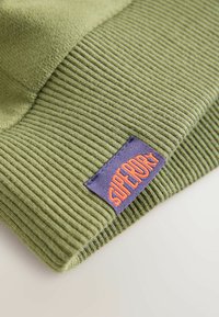 Zelená pletená čepice s ribkovou strukturou, s fialovým štítkem s nápisem "SUPERDRY" v oranžové barvě. Tkanina má měkký a teplý vzhled.