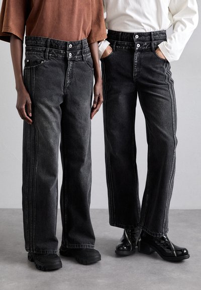 Jean en denim noir à jambes larges avec fermetures à double bouton, rayures latérales et poches avant, associés à des chaussures noires.