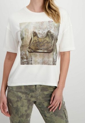 Kobieta w białej koszulce z nadrukiem beżowej torebki z łańcuszkiem, w połączeniu z zielonymi spodniami w kamuflażowy wzór.