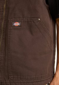 Brown canvas vest featuring a front zipper, two side pockets, and a logo patch on the left chest. Textured fabric with durable stitching.