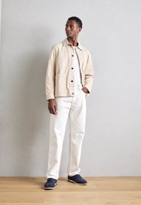 Beige canvasjacka med knappframsida, vit t-shirt, vita byxor och navy mockaskor. Står på ett trägolv mot en grå vägg.