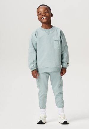 Hellblaues Sweatshirt mit Brusttasche und passenden tapered Pants. Weiße Sneakers mit schwarzen Akzenten. Glatter Stoff und lässige Passform.