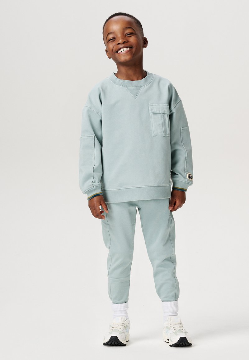 Hellblaues Sweatshirt mit Brusttasche und passenden tapered Pants. Weiße Sneakers mit schwarzen Akzenten. Glatter Stoff und lässige Passform.