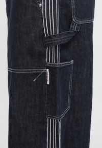 Dunkelblaue Jeans-Latzhose mit weißen Nähten, mehreren Taschen und einem pinkfarbenen Logo-Tag. Entworfen mit geradem Schnitt und strapazierfähiger Stoffstruktur.