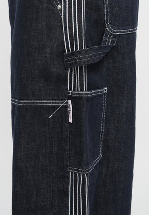 Dunkelblaue Jeans-Latzhose mit weißen Nähten, mehreren Taschen und einem pinkfarbenen Logo-Tag. Entworfen mit geradem Schnitt und strapazierfähiger Stoffstruktur.