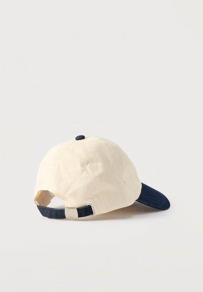 Casquette en tissu beige avec une visière bleu marine foncé. Dispose d'une sangle ajustable avec un accent métallique à l'arrière. Texture douce et forme arrondie.