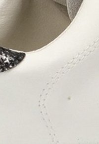 Zapato blanco de cuero con un acento negro texturizado cerca de la abertura. Superficie lisa con costuras visibles e imperfecciones menores.