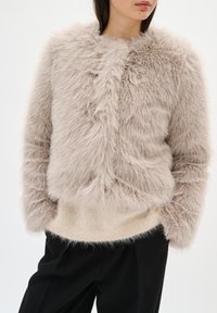 Chaqueta de pelo sintético en beige claro, diseño corto con textura mullida, dobladillo acanalado y hombros caídos, combinada con un suéter beige suave y pantalones negros.