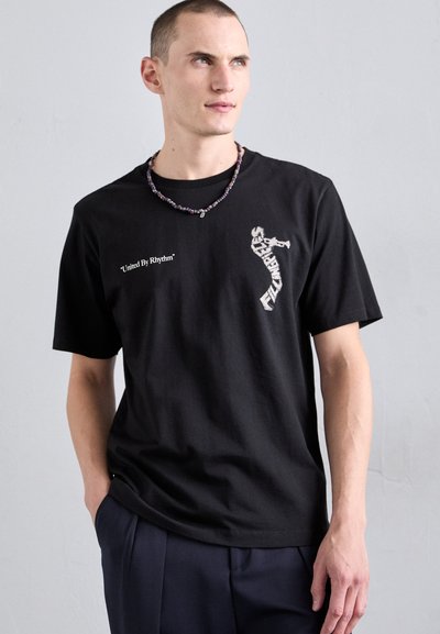 Svart bomullströja med korta ärmar, prydd med vit text "United By Rhythm" och en grafik på framsidan. Modellen bär ett flerdelat pärlhalsband.