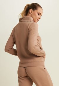 Giacca zip-up marrone chiaro con colletto alto e texture. Tessuto morbido, design aderente e tasche laterali. Indossata con pantaloni coordinati a fantasia.
