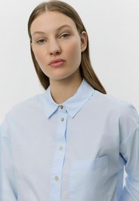 Cămașă ușor albastră, din bumbac. Are un guler clasic, buzunar frontal și accente de butoni bej. Textură netedă.